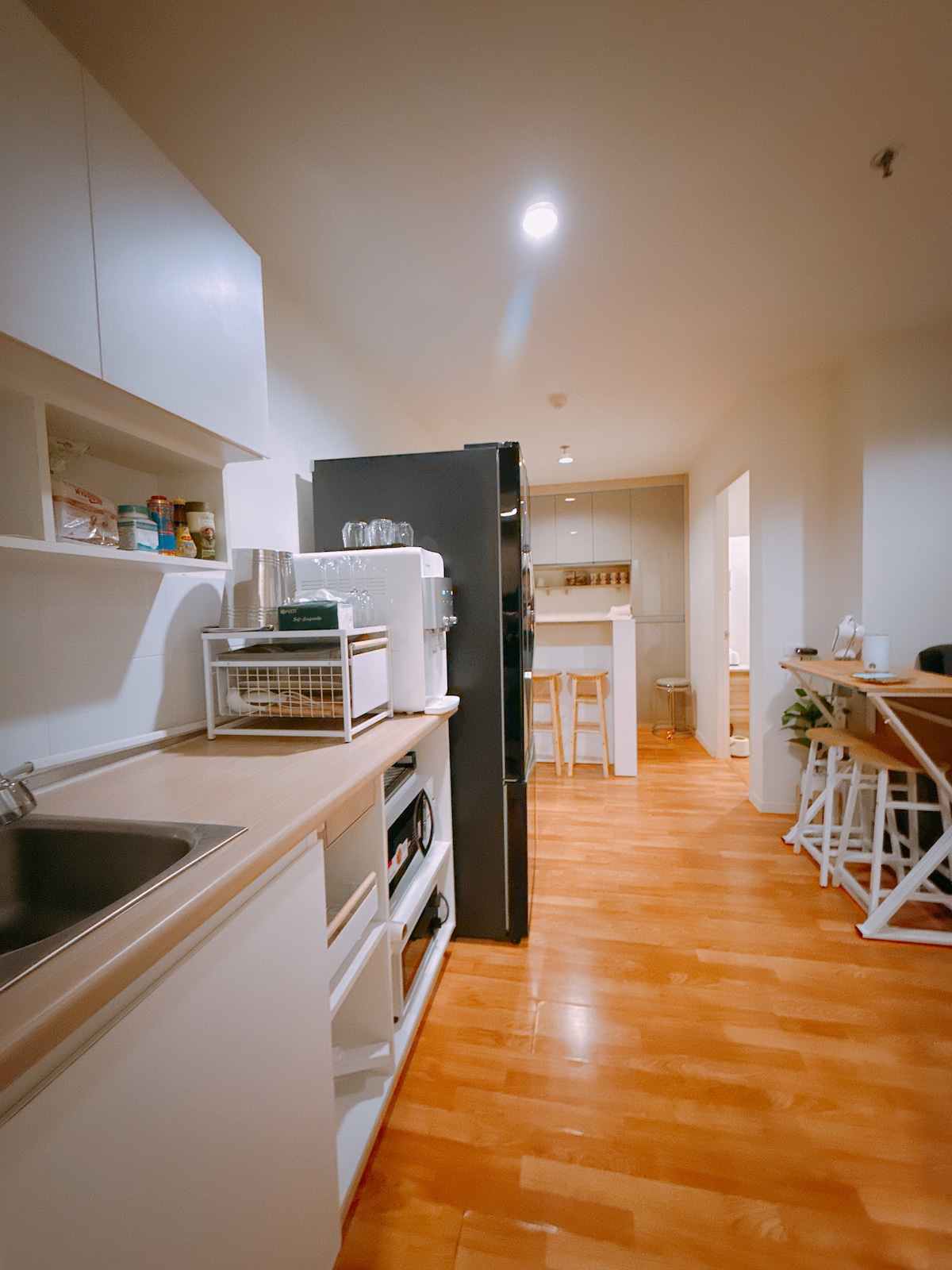 A clean and functional kitchen features white cabinetry and wooden countertops. A stainless steel sink is positioned beside a compact refrigerator. The space is illuminated by soft ceiling lights, with a cozy dining area visible in the background, enhancing accessibility for meal preparation.