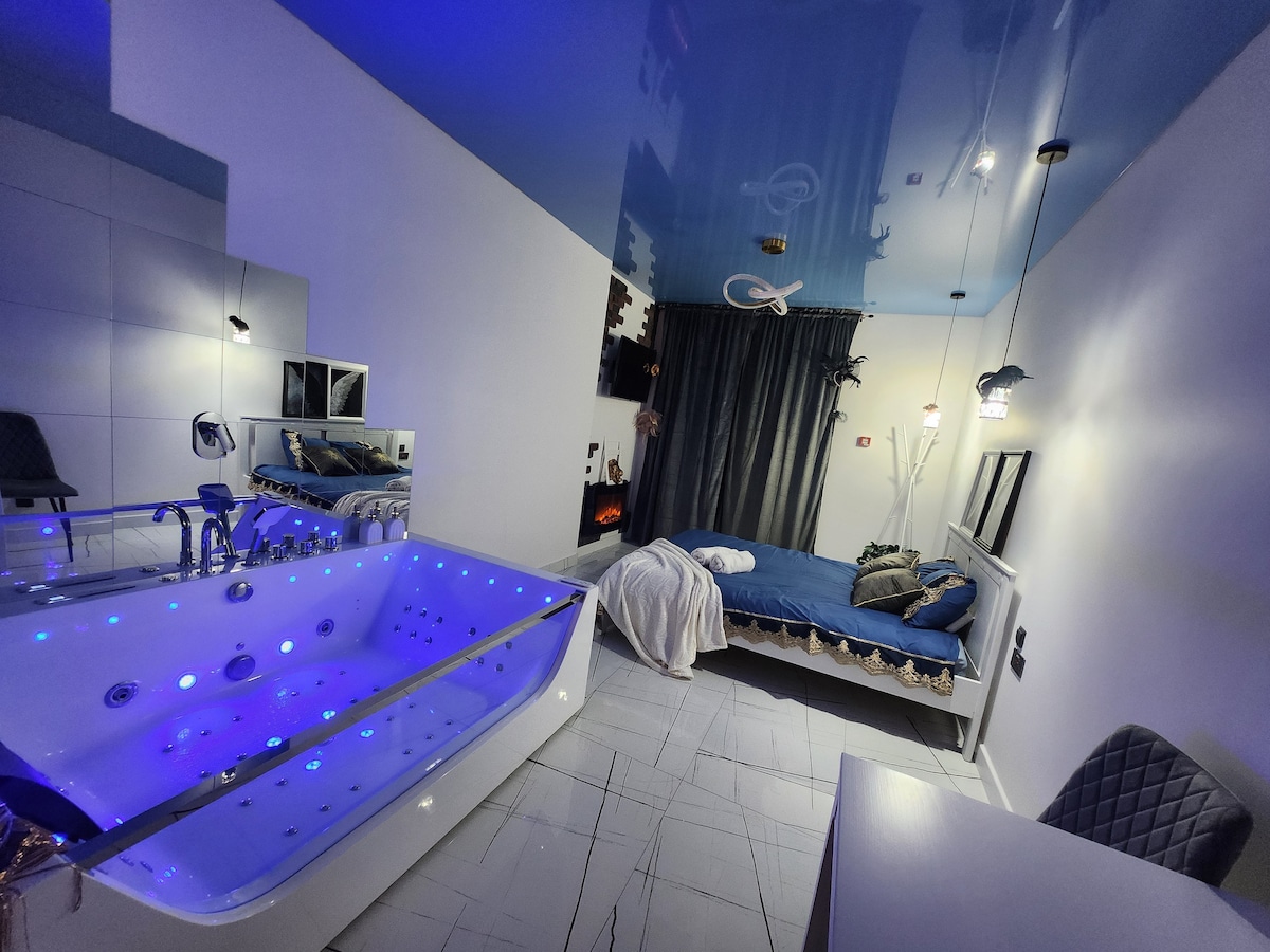 A modern interior showcases a spacious jacuzzi illuminated with blue lights, paired with a cozy sleeping area featuring a double bed. A stylish desk and a large mirror add functionality to the space. Dark curtains frame the window, creating a serene atmosphere.