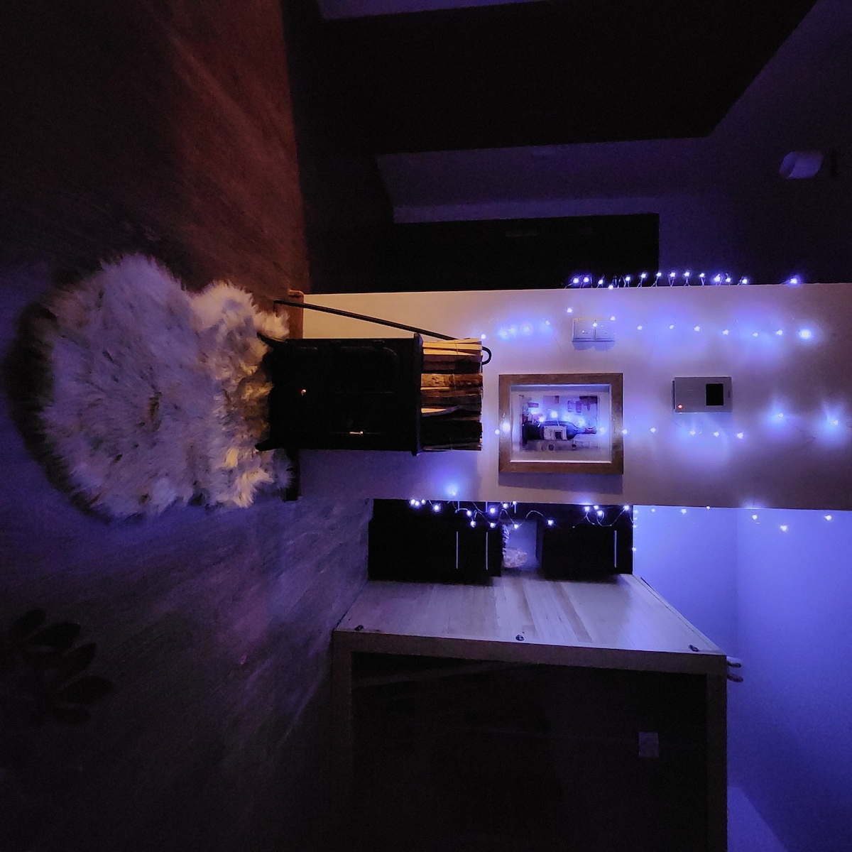 A cozy interior space is highlighted by soft, dim lighting from string lights along the wall. A small cart is positioned to the side, adorned with rolled towels. A light-colored rug with a fluffy texture is positioned on the floor, adding warmth to the ambiance.