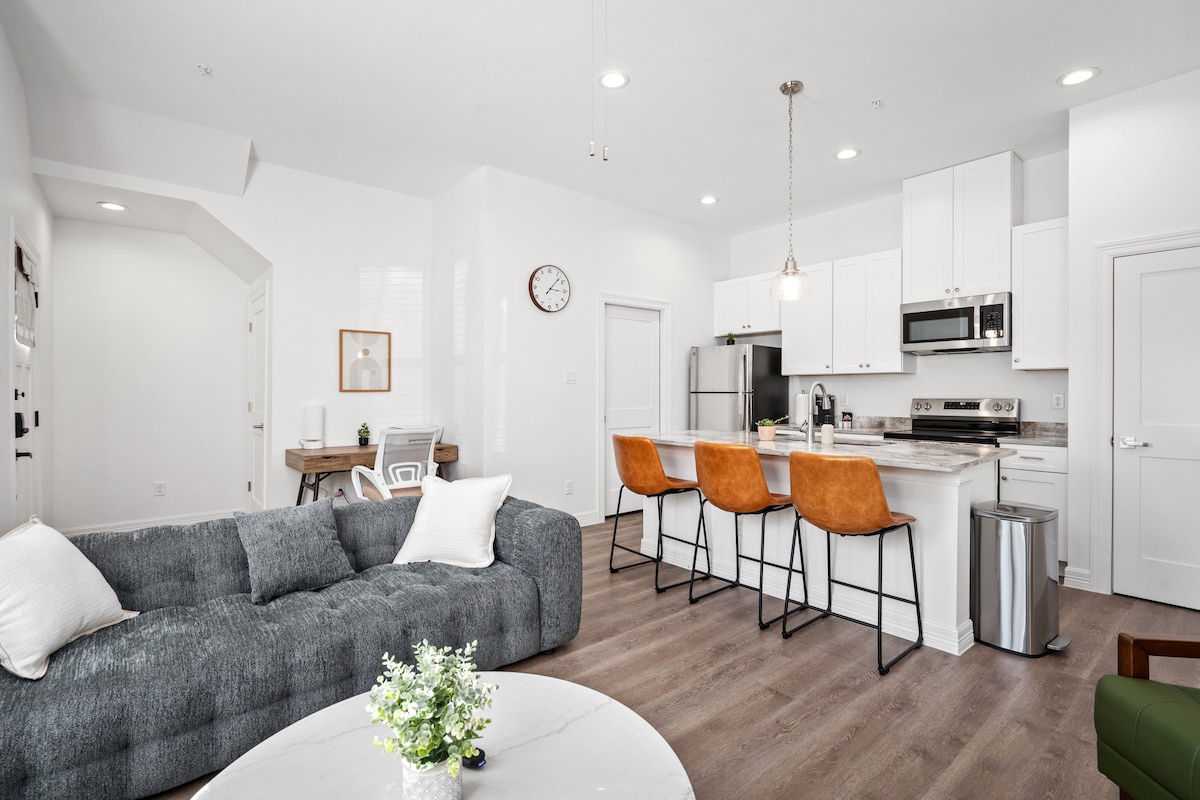 An open living space features modern furnishings, including a comfortable gray sofa and a round coffee table adorned with a small plant. The kitchen showcases sleek white cabinets, stainless steel appliances, and three orange bar stools at the counter. Soft lighting enhances the inviting atmosphere.