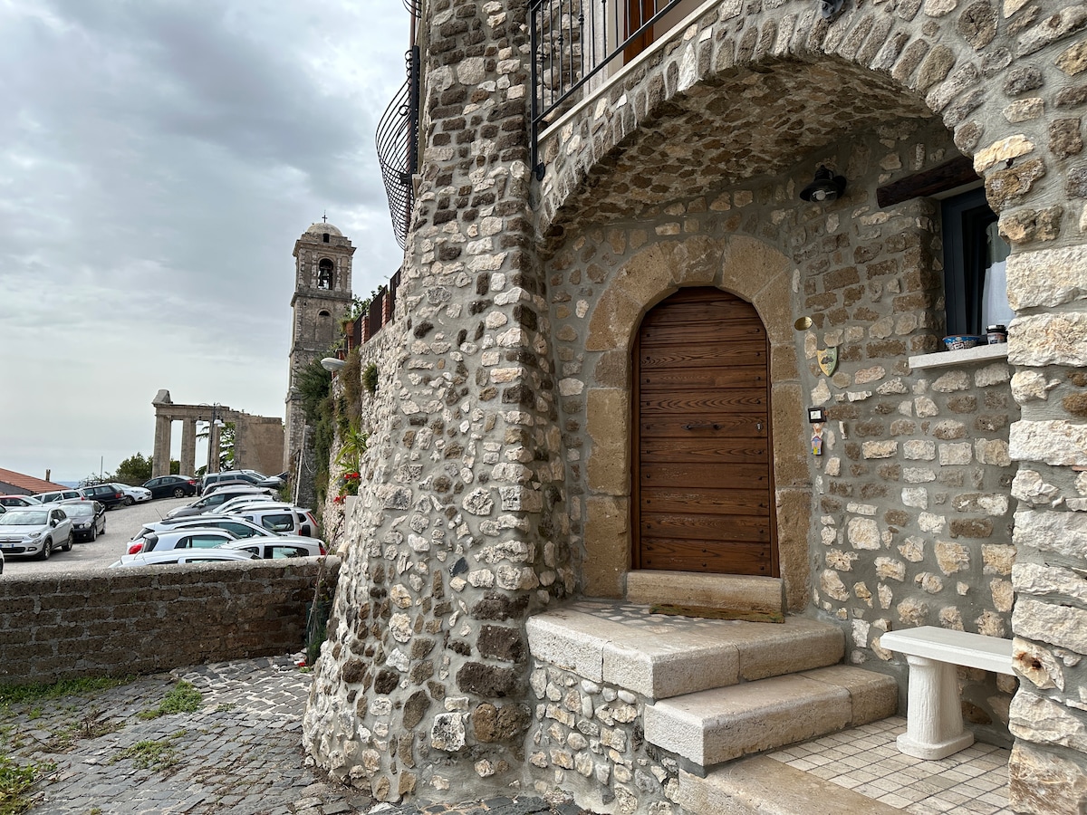 A stone building is showcased, featuring a large wooden door that stands out against the textured wall. The entrance is framed by smooth stone steps and a small seating area. In the background, a bell tower and ancient ruins are visible, enhancing the historical ambiance.