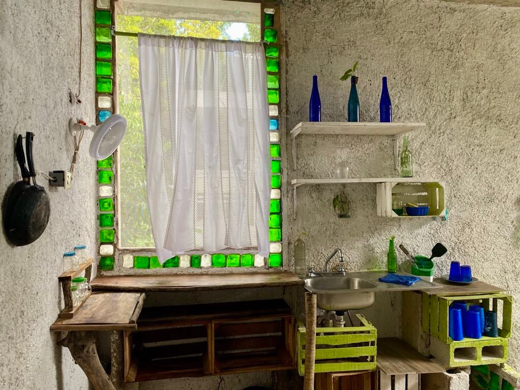 A unique kitchen area features a rustic wooden counter and shelves made from repurposed materials. Colorful green and blue glass bottles are displayed near a window with a sheer curtain, allowing natural light to filter through, complementing the textured walls.