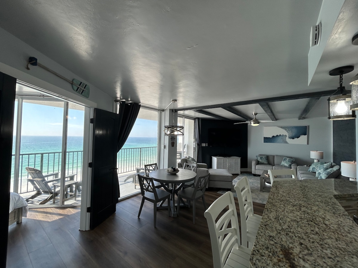 An open living area features a dining table with white chairs and a kitchen counter. Large windows showcase a view of the ocean, with natural light streaming in. Comfortable seating is arranged nearby, creating a welcoming space with beautiful coastal decor.