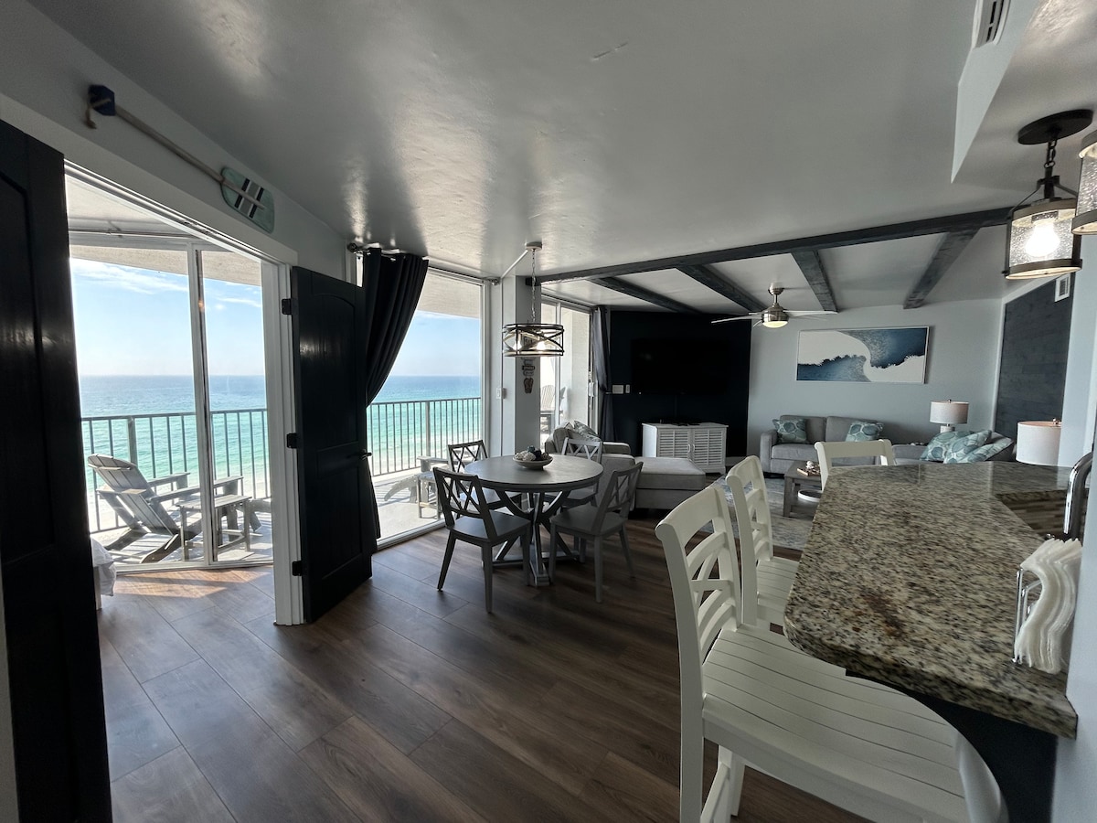 An open-concept living area features a dining table surrounded by light-colored chairs, with panoramic ocean views visible through large glass doors. A well-appointed kitchen with a granite countertop is in the foreground, while comfortable seating is arranged in the inviting living space beyond.