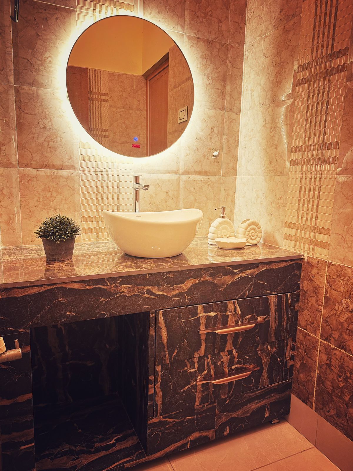 The bathroom features elegant marble accents with a sleek, modern design. A round mirror framed with soft lighting illuminates a stylish sink atop a dark, patterned vanity. Simple decor elements, including a small plant, add a touch of warmth to the space.
