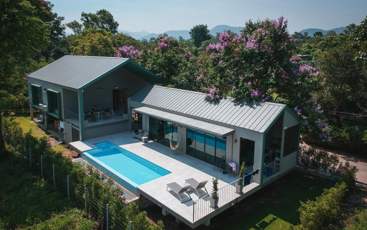 An aerial view captures the modern home surrounded by greenery, featuring a sleek saltwater pool situated beside a spacious patio with loungers. Lush trees and blooming flowers complement the outdoor tranquility, while mountains are visible in the background under a clear blue sky.