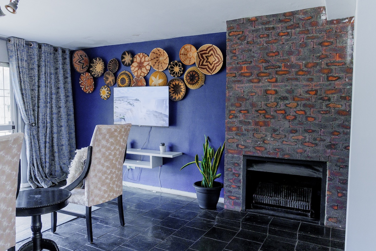 A stylish living area features a large wall adorned with decorative wooden plates. A flat-screen television is mounted on the wall opposite a cozy fireplace. Comfortable chairs and a small table create a welcoming space, complemented by a potted plant in the corner.
