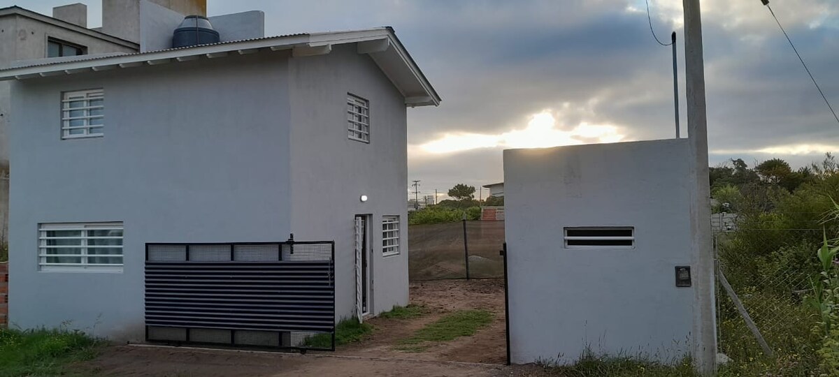 A modern two-story building is depicted with a light gray exterior. Lattice windows provide natural light, while a black gated entrance leads to a fenced outdoor area. Soft sunlight is seen peeking through the clouds in the background, creating a serene environment.