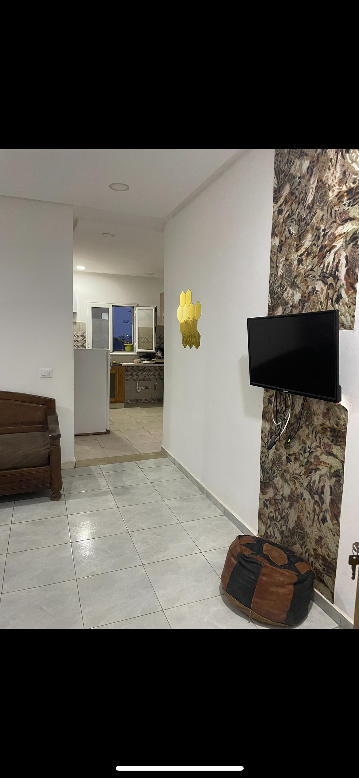 A hallway is shown leading to the kitchen, with light-colored tiles and white walls. A compact television is mounted on a feature wall with a textured design, while a brown bag and a wooden chair are placed on the floor, adding functional elements to the space.