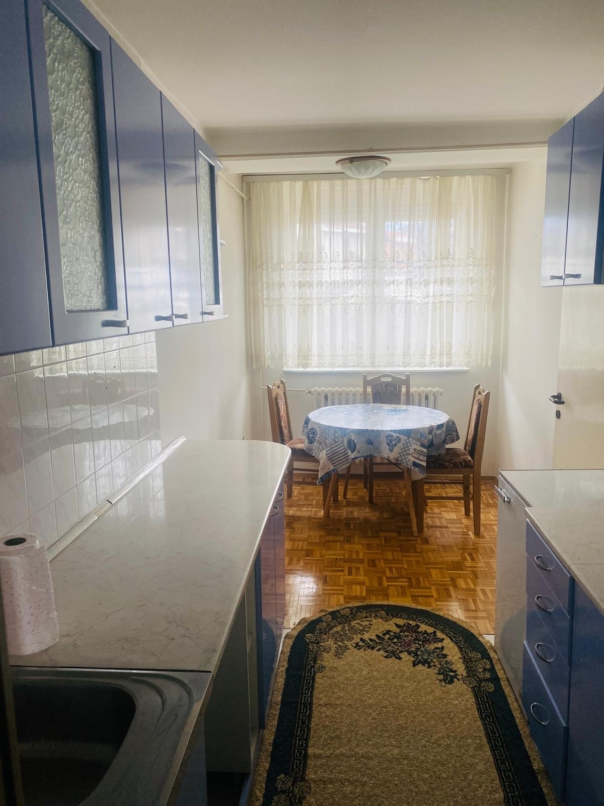 A compact kitchen area features light blue cabinetry and a countertop. Natural light filters through sheer curtains, illuminating a small dining table set with a blue tablecloth and surrounded by four chairs. A patterned rug adds warmth to the space.