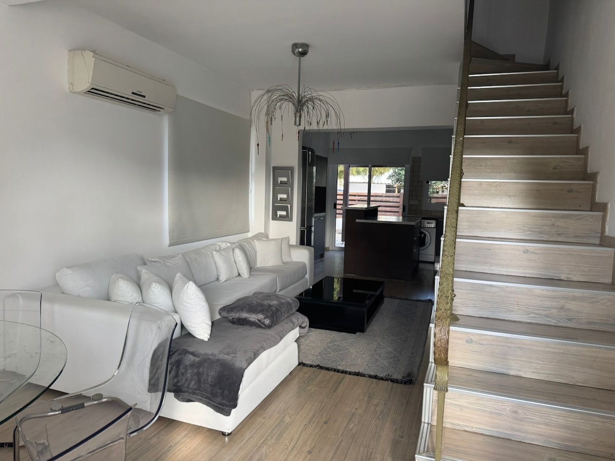 A light-filled living area features a comfortable white sofa adorned with fluffy pillows, complemented by a glass coffee table. A stylish staircase ascends to the upper level, while glass doors provide a view of the outdoor space.