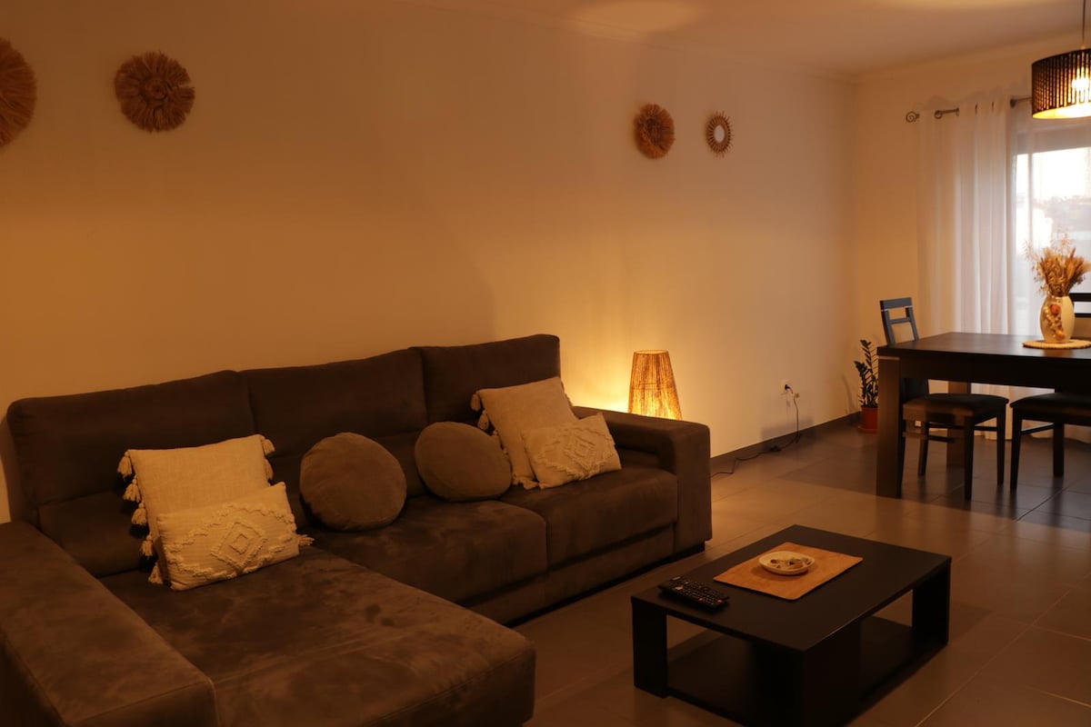 A modern living area features a sectional sofa adorned with decorative cushions. A coffee table sits in front, accompanied by a dining table positioned adjacent to a window, where soft light enters, illuminating the space. Warm accents enhance a relaxed atmosphere.