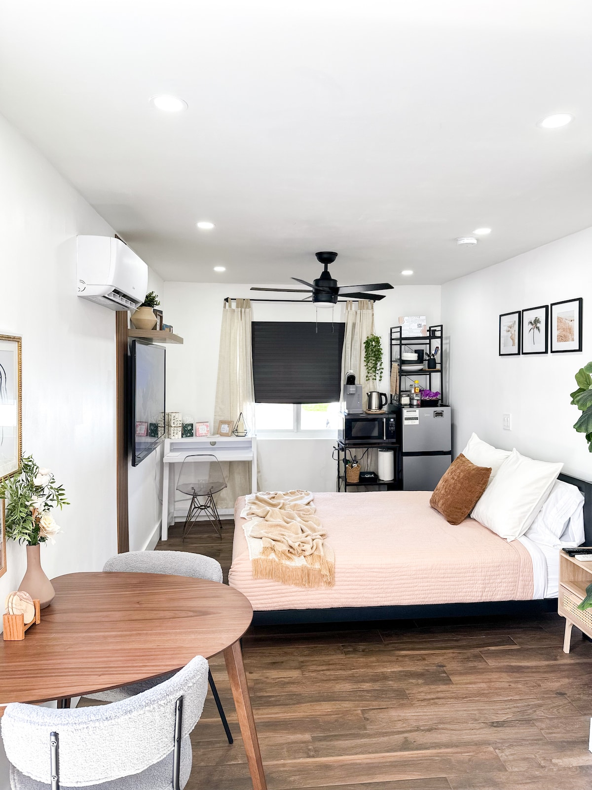 A cozy studio space features a comfortable queen bed dressed in soft linens, complemented by a decorative throw. A small round dining table is situated in the foreground, while a kitchenette with a mini fridge and microwave is positioned in the background, alongside a dedicated workspace.