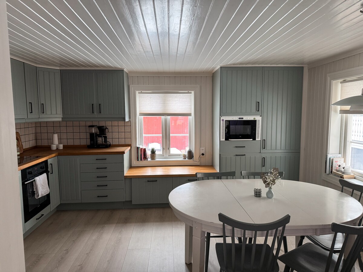 A well-equipped kitchen features light green cabinetry and a wooden countertop. Two windows allow natural light to illuminate the space, while a round dining table with black chairs offers seating for several guests. Appliances include an oven, microwave, and coffee maker, ensuring functionality.