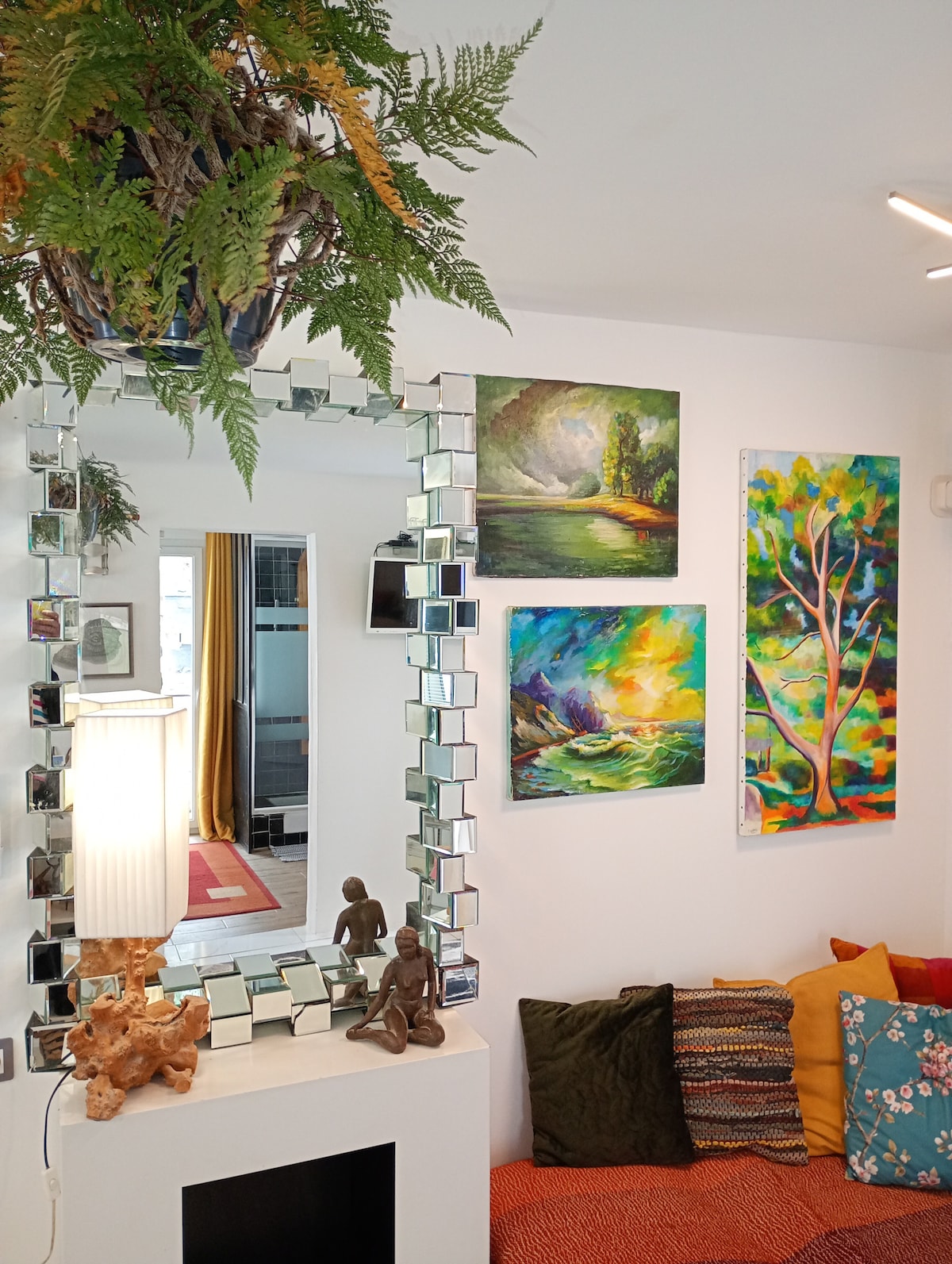 An inviting living area features a mirror framed with small square tiles. Colorful artwork adorns the walls, showcasing various landscapes and nature scenes. A cozy seating arrangement with patterned cushions is positioned near a decorative sculpture, enhancing the welcoming ambiance of the space.