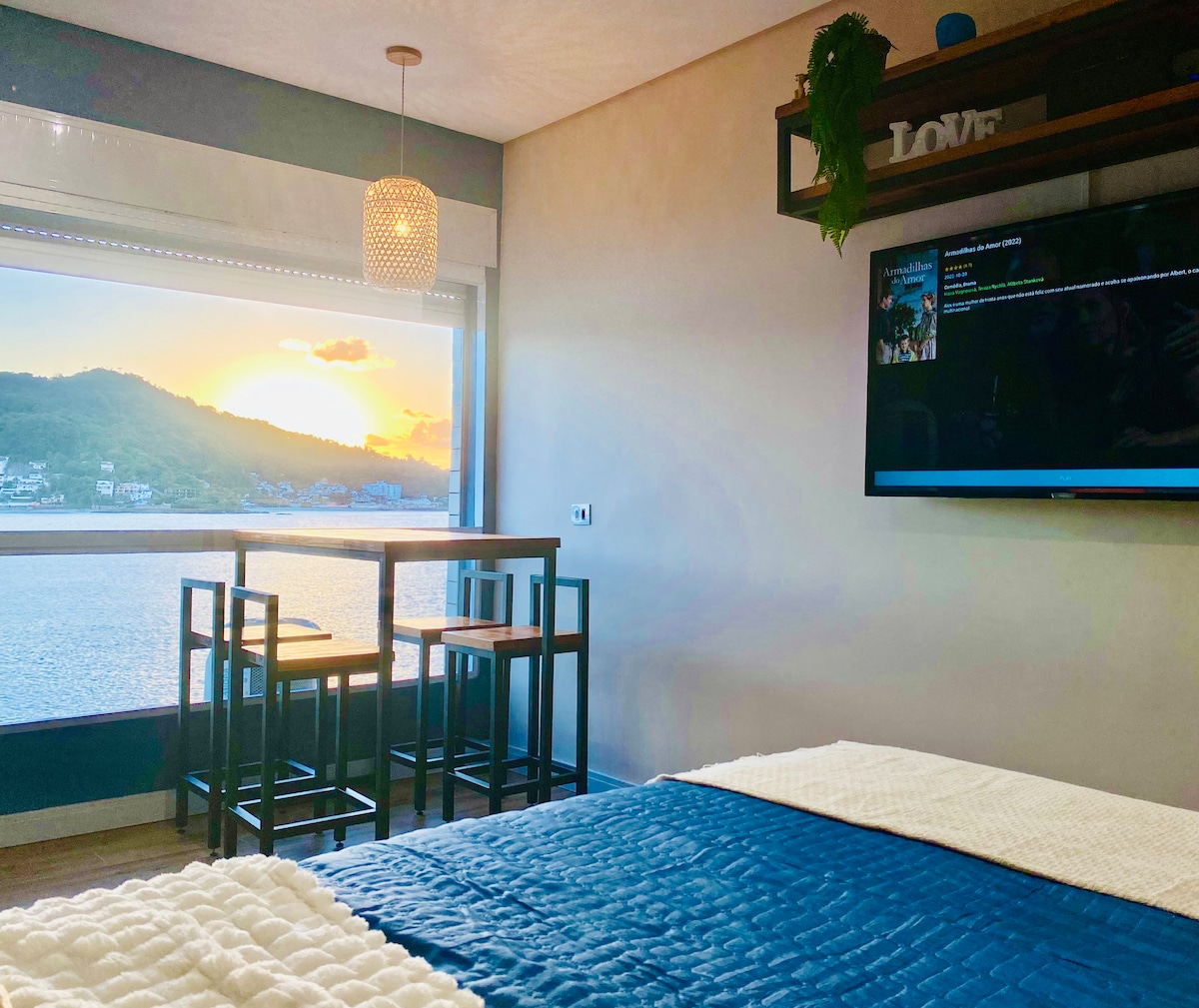 The room features a cozy bed with a textured blue coverlet. A wall-mounted television is positioned opposite, with a dining table and stools offering a view of the sunset over the ocean through a large window.