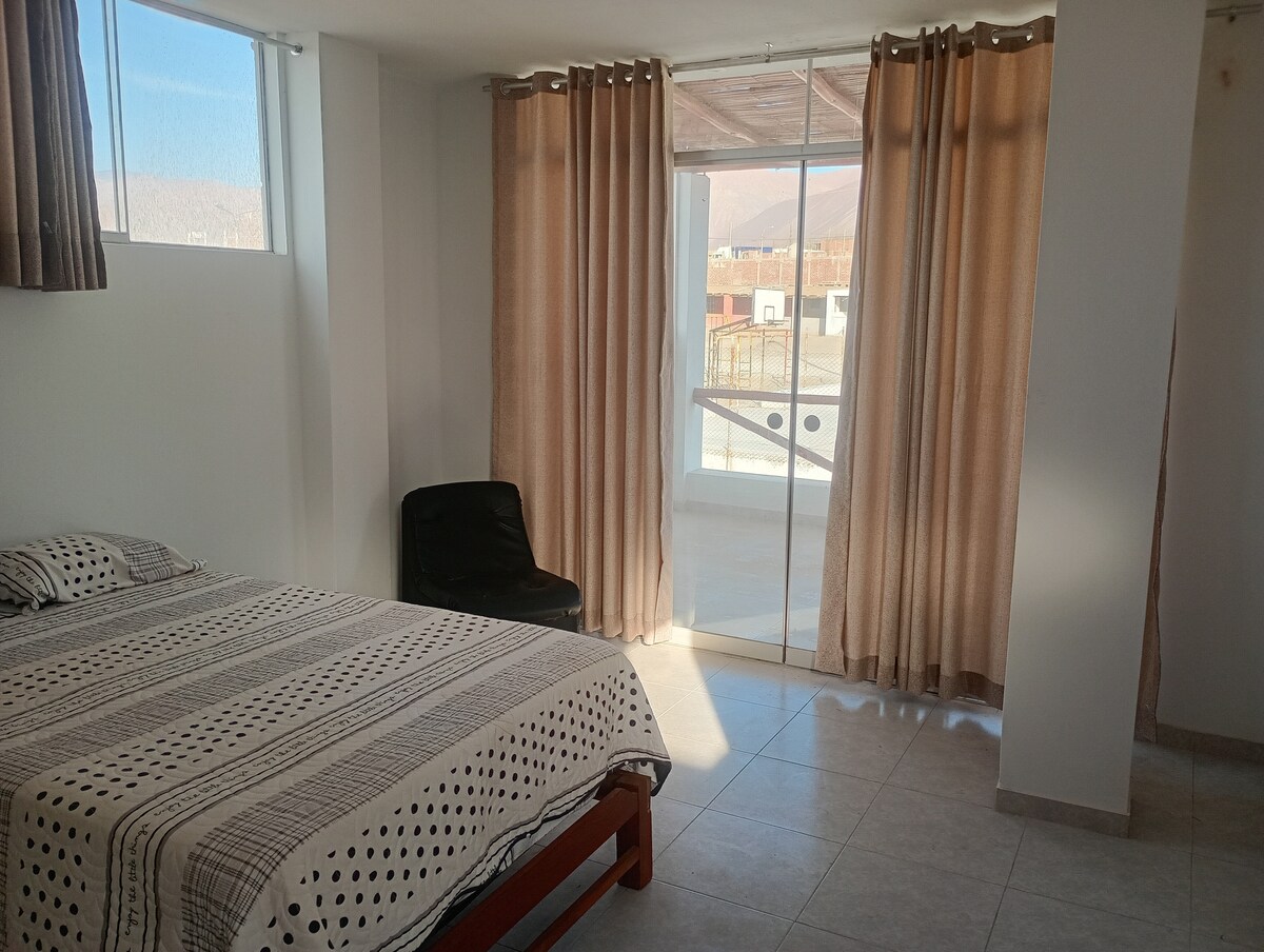 A bright sleeping area features a spacious bed with a patterned blanket. Natural light floods through large windows, complemented by sheer curtains. A simple chair is positioned nearby, and a sliding glass door provides access to a private outdoor space, enhancing the room's openness.