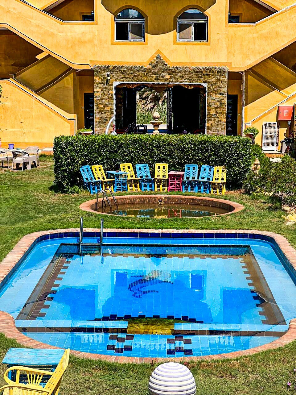 A serene outdoor space features a clear blue pool surrounded by vibrant chairs in various colors. Lush greenery frames the area, and a small hot tub is visible adjacent to the pool. The backdrop consists of a distinctive yellow building with large windows.