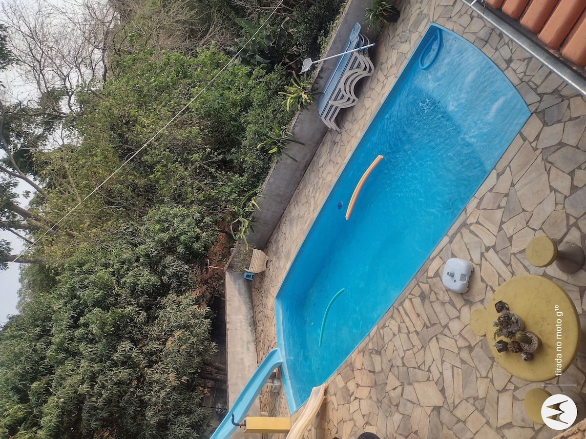 A clear swimming pool is surrounded by a stone patio, featuring a small table and chairs. Lush greenery lines the perimeter of the area, providing a natural backdrop. A poolside slide adds an element of fun to the space, enhancing its appeal for family enjoyment.