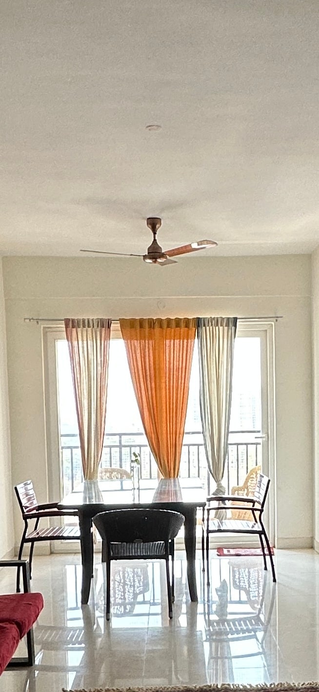 A dining area is visible with a simple black table surrounded by four chairs. Sheer curtains in varying shades of orange and grey frame a large window, allowing natural light to fill the space. A ceiling fan is positioned above the table.