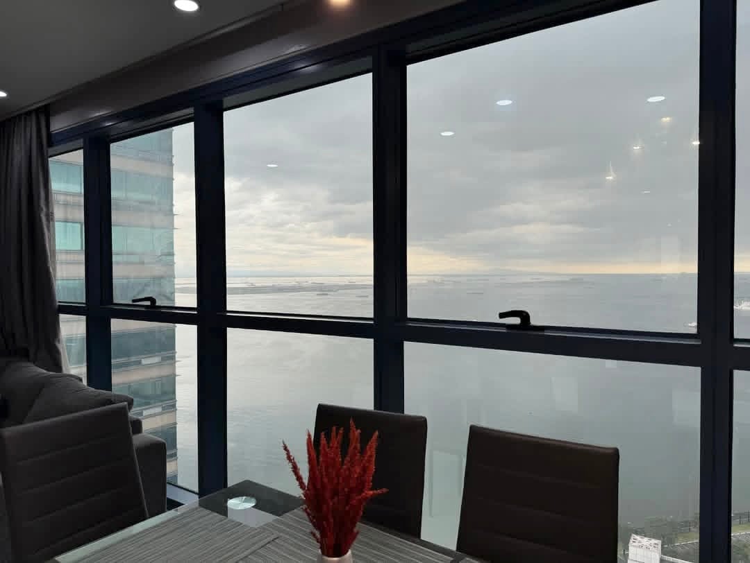 Large windows offer expansive views of Manila Bay against a cloudy sky, allowing natural light to fill the interior space. A dining table with dark chairs and a centerpiece sits in the foreground, enhancing the inviting atmosphere of the room.