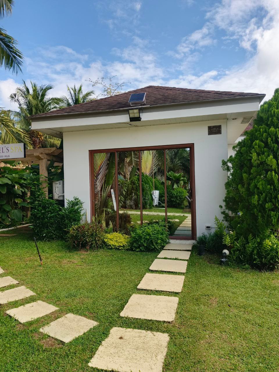 A charming spa structure is framed by natural greenery, featuring large glass doors that reflect the surrounding landscape. A pathway of stone pavers leads to the entrance, enhancing the inviting garden setting under a clear sky.
