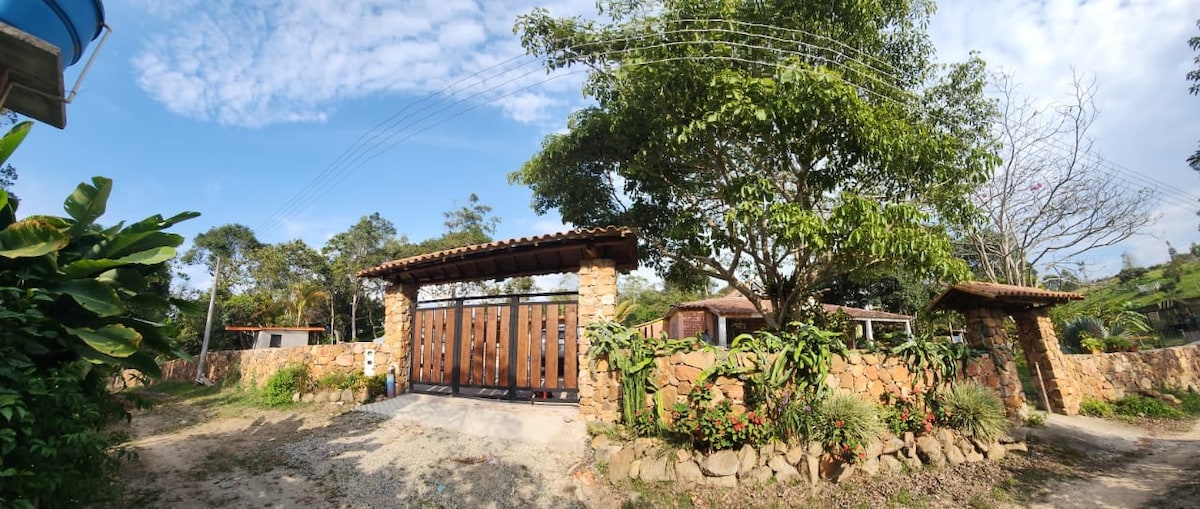The entrance to the property features a rustic stone wall adorned with lush greenery and vibrant flowers. A pair of wooden gates stands open, inviting guests into the serene environment. A clear blue sky complements the natural setting, enhancing the peaceful ambiance.