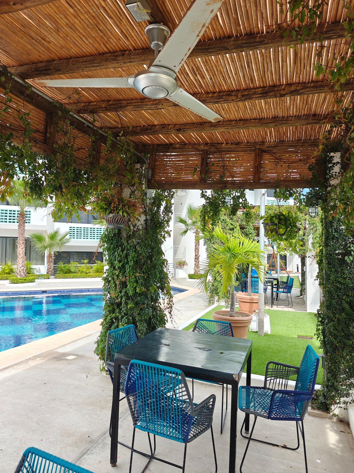 An outdoor seating area is framed by lush greenery, featuring a shaded table with four blue chairs. The scene is complemented by a clear swimming pool in the background, surrounded by palm trees and well-maintained landscaping.