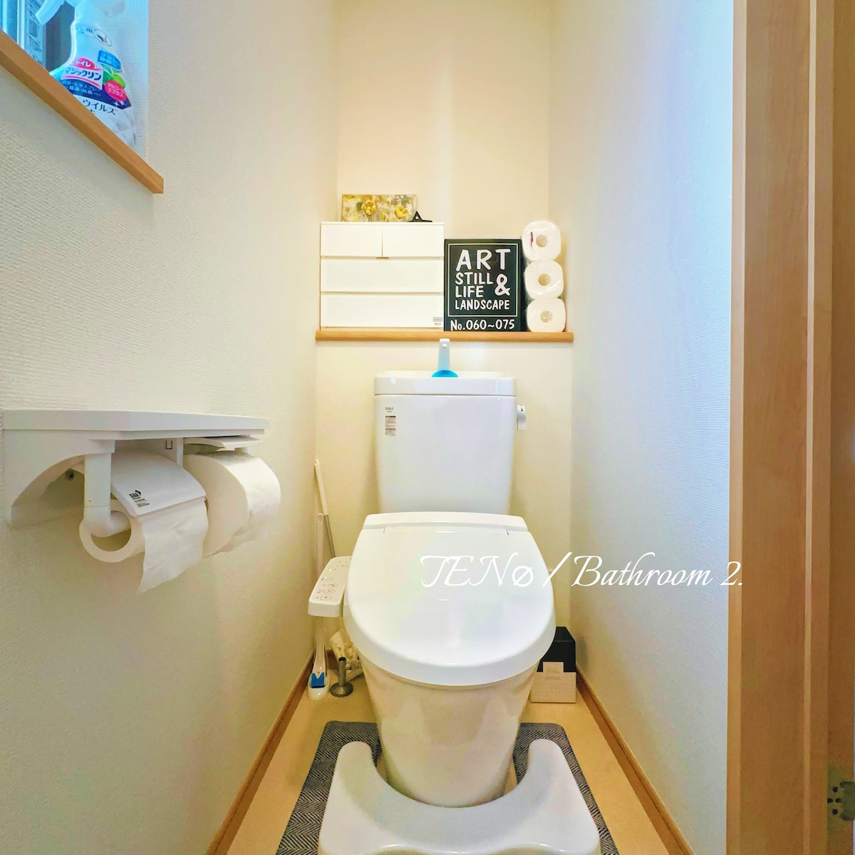 A clean and functional bathroom is presented, featuring a modern toilet with a soft-close seat. Wall-mounted shelves hold decorative items and toiletries. A roll of toilet paper is easily accessible, and the floor includes a small mat in front of the toilet for comfort.