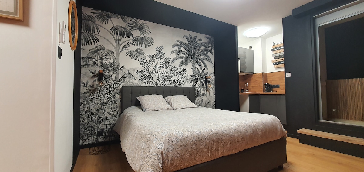 A spacious sleeping area is highlighted by a decorative wall featuring a tropical motif. The large bed, dressed in a light patterned coverlet, is situated against the wall. A kitchenette area is visible in the background, complemented by warm wood accents.