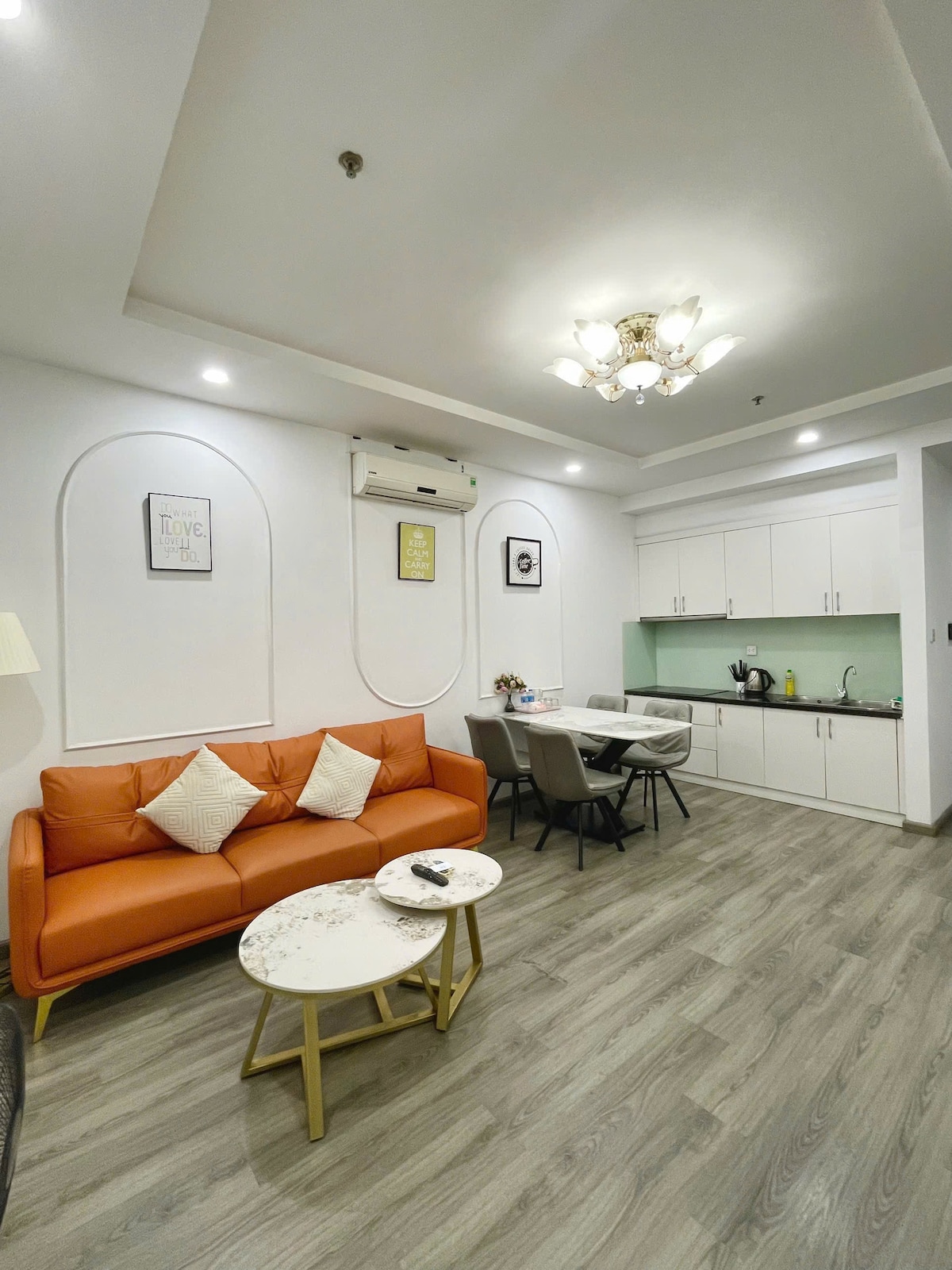 A modern living area is showcased with an orange sofa and two decorative pillows. A round coffee table sits on a wood-like floor, adjacent to a dining table surrounded by chairs. A well-equipped kitchenette is visible, featuring white cabinetry and a green backsplash.