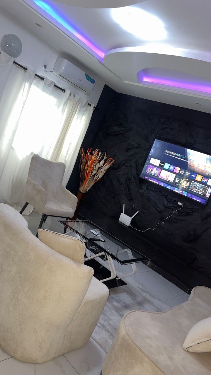 A modern living area is presented, featuring light-colored upholstered chairs set around a glass coffee table. A large television is mounted on a textured wall, and soft light from the windows is complemented by a subtle ceiling hue. An artistic plant adds a touch of color.
