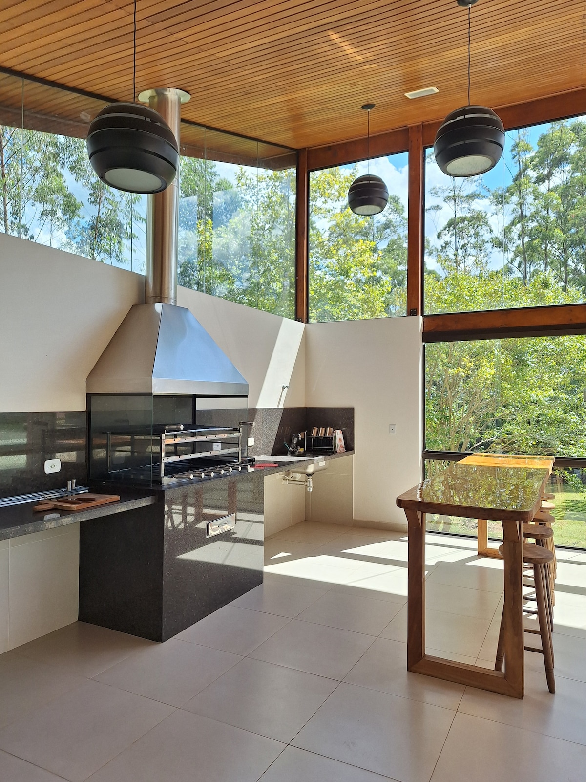 A bright kitchen features large windows that bring in natural light and offer views of the surrounding trees. A sleek countertop is equipped with modern appliances, including an oven and cooktop. A wooden bar with stools provides a casual dining option.