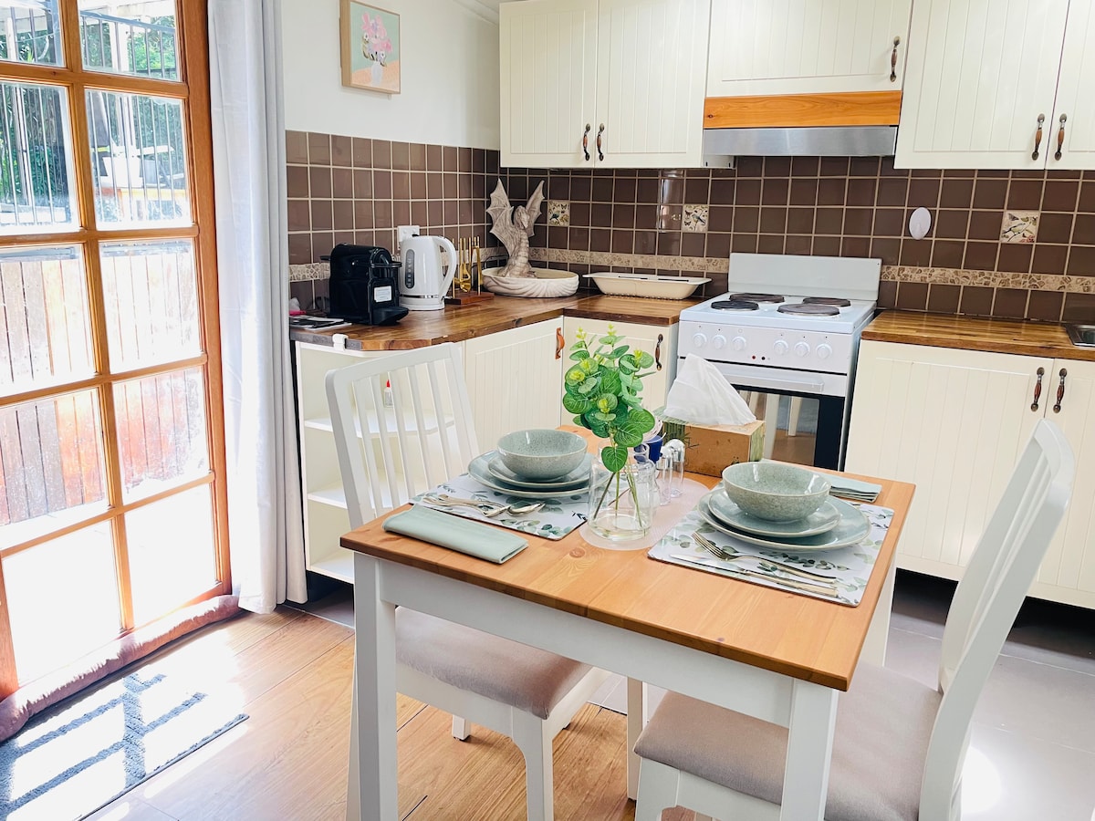 A bright kitchen space features a wooden dining table set for two, surrounded by white chairs. Natural light floods in through glass doors, highlighting the tiled backsplash and wooden cabinetry. Essential appliances, including a kettle and stove, are neatly arranged for convenience.