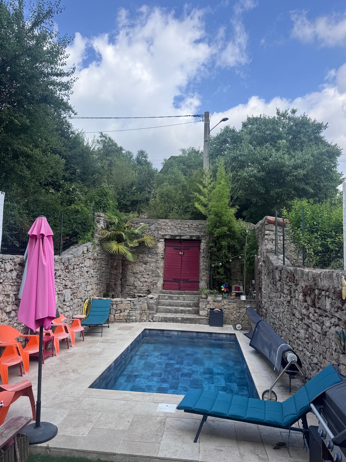 A shared outdoor space is highlighted by a swimming pool surrounded by various seating options, including colorful lounge chairs. Natural greenery, stone walls, and a decorative gate provide a rustic backdrop against a partly cloudy sky.