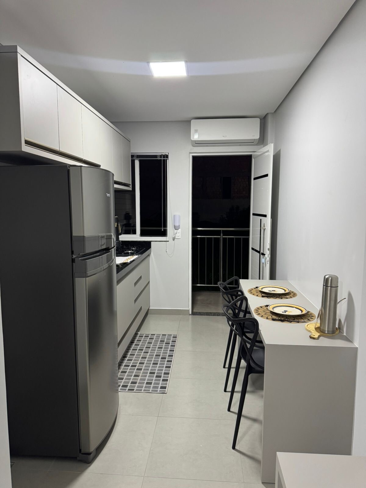 A modern kitchen is equipped with stainless steel appliances, including a refrigerator and stove. A small dining area is set for two, featuring placemats on a white table. Natural light filters in through the window, which overlooks a balcony.
