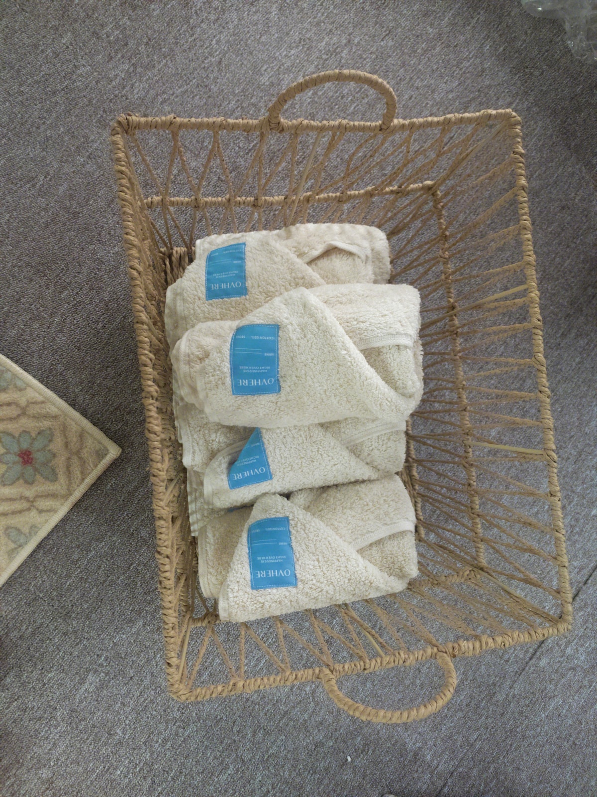 A woven basket holds neatly folded towels, presenting a soft, neutral colour with a tag visible on each. The towels are arranged uniformly, offering a tidy appearance. A patterned rug is visible in the lower corner, adding texture to the setting.
