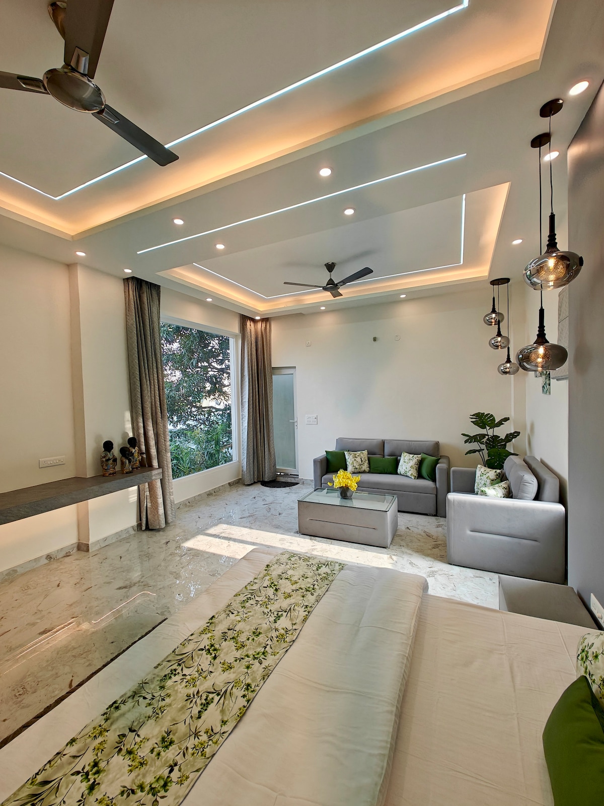 A spacious studio apartment features modern decor with a bed adorned with floral bedding. A cozy sitting area includes a 2-seater sofa and a coffee table. Abundant natural light enters through a large window, complemented by soft, ambient ceiling lighting.