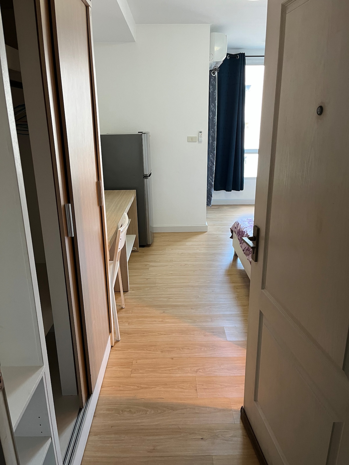 A narrow hallway leads to a well-lit living area furnished with a small table and a chair. The space includes a refrigerator and features wooden flooring, creating a simple and functional atmosphere. A window provides natural light, and dark curtains partially frame it.