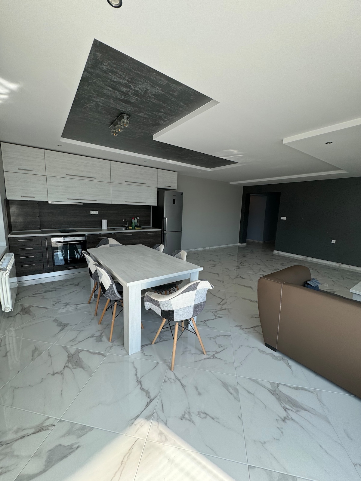 An open-concept living area is displayed, featuring a dining table surrounded by stylish chairs. A modern kitchen with integrated appliances is visible in the background, complemented by sleek cabinetry. Glossy tile flooring enhances the spacious ambiance, and natural light fills the room through large windows.