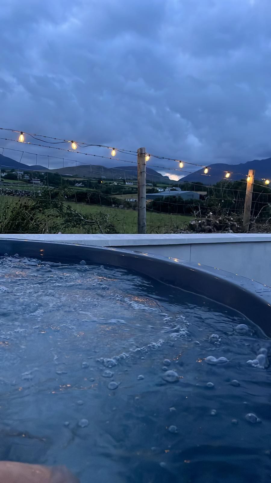 A hot tub is shown with bubbles rising to the surface against a backdrop of distant mountains. Outdoor string lights are visible overhead, illuminating the space softly as evening approaches. Lush greenery is present in the landscape surrounding the area.