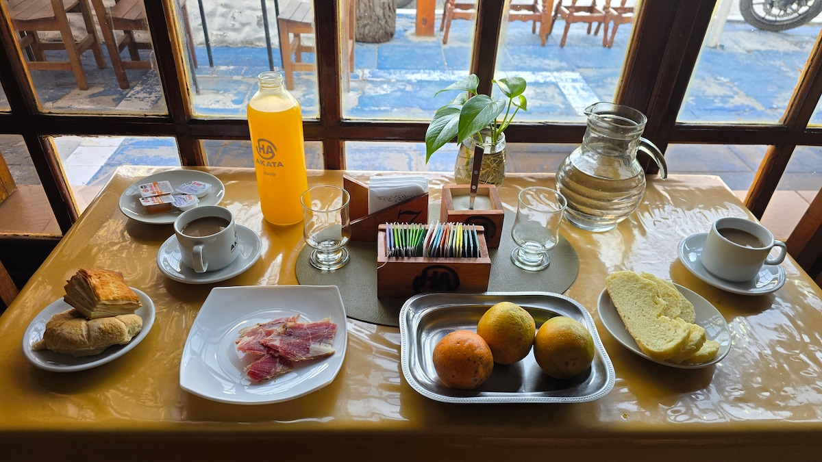 A breakfast table is set with a variety of items. Two cups of coffee sit next to an orange juice pitcher. A selection of pastries, meats, fruits, and bread is arranged neatly on plates, with a small plant adding a touch of greenery to the inviting scene.