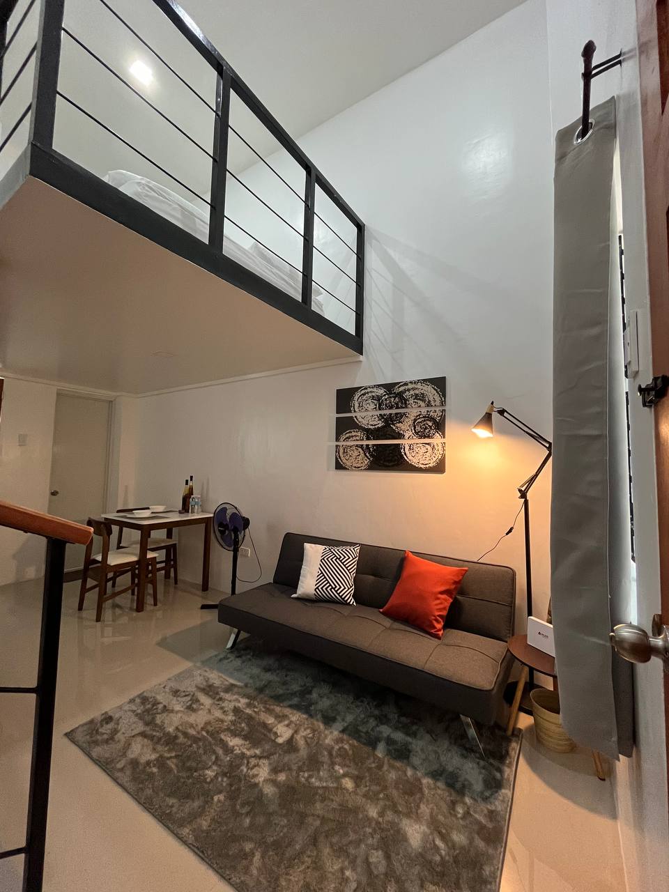 A modern loft living area is shown, featuring a gray sofa accented with orange and black pillows. A rug lies underneath, complemented by a standing lamp. In the background, a small dining table and chairs are visible, with a staircase leading to the loft bedroom above.