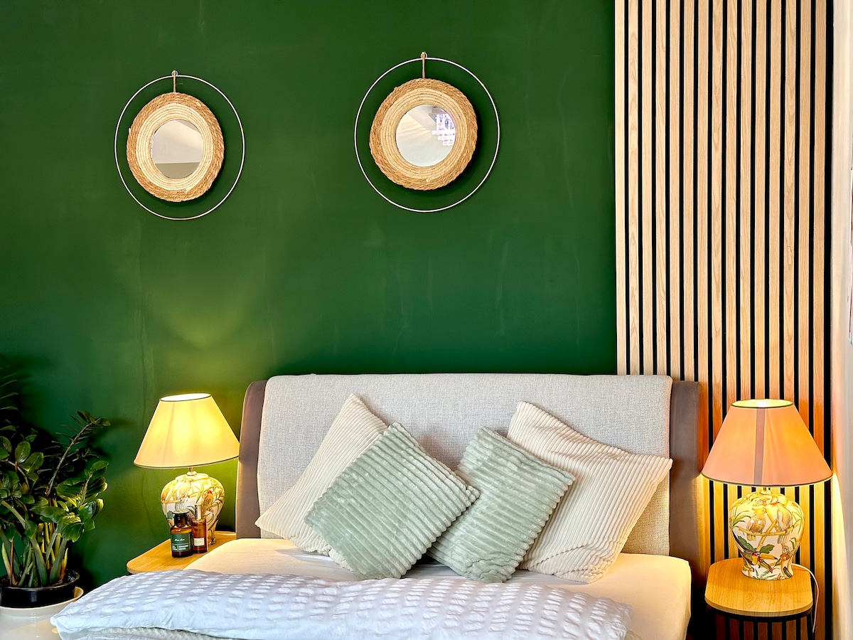 A cozy bedroom features a green accent wall adorned with two round mirrors. A spacious bed is complemented by decorative pillows in soft hues. On either side of the bed, bedside tables hold lamps that provide warm lighting, creating an inviting atmosphere.
