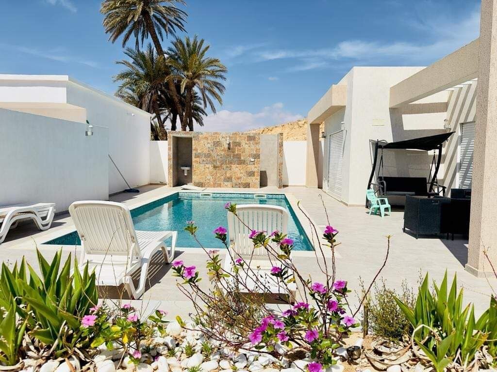 A private pool area features a rectangular pool surrounded by lounge chairs. Lush greenery and colorful flowers line the space, while a stone wall and palm trees provide a serene backdrop. A shaded seating area is visible to the side, enhancing the relaxation setting.