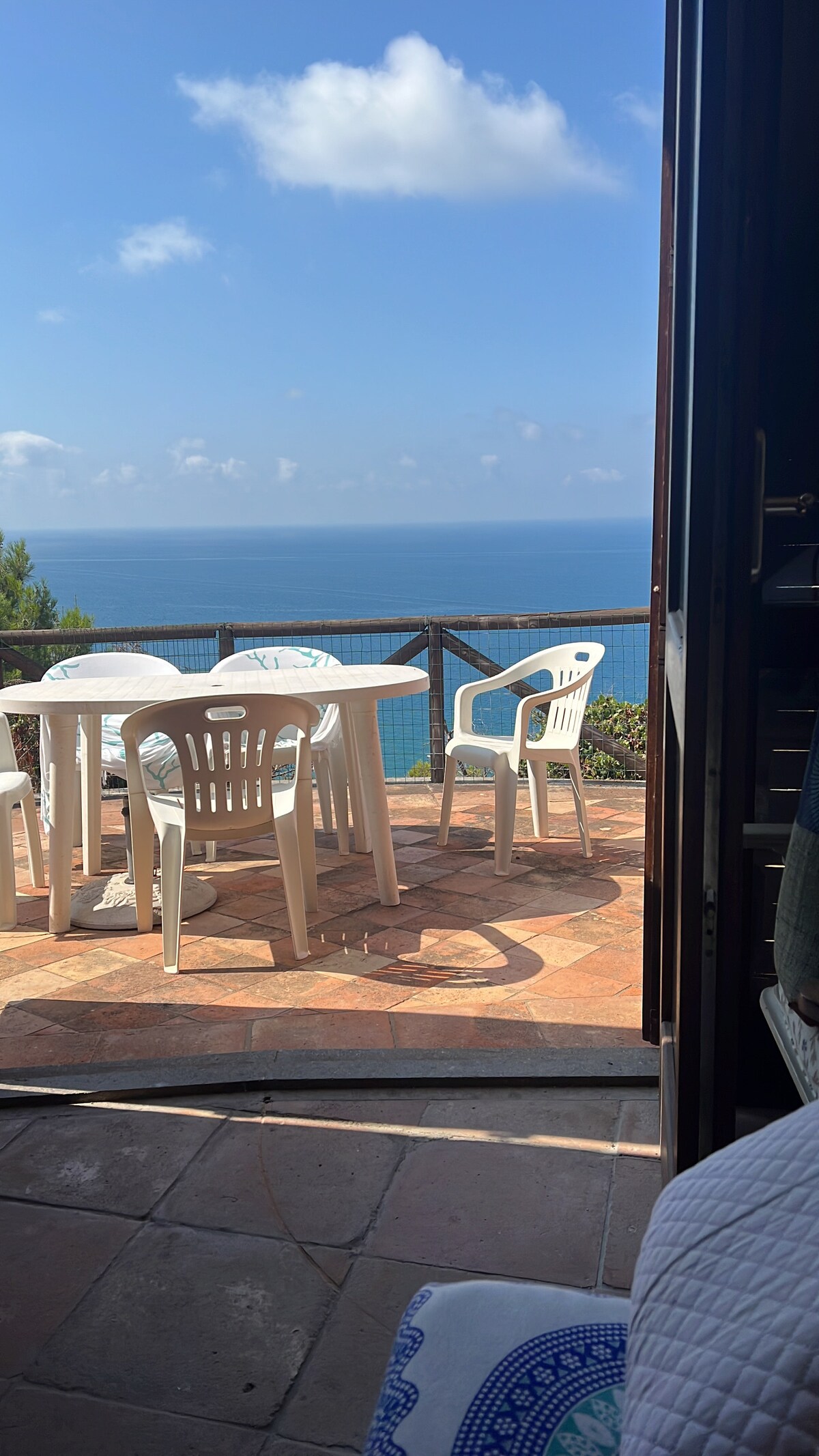 A terrace with a table and several white chairs overlooks the sea, framed by a doorway. The sunlit space offers expansive ocean views, with a clear blue sky and scattered clouds above. The tiled flooring complements the peaceful outdoor environment.