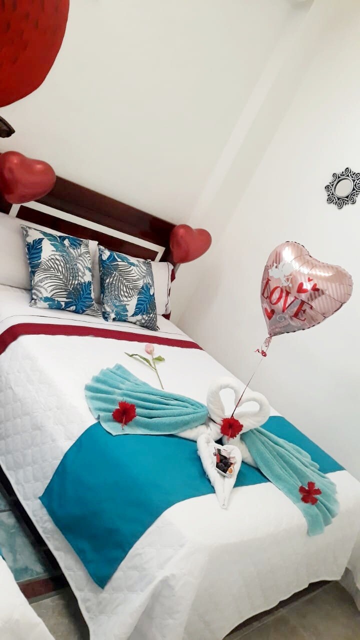 A spacious bed is adorned with a white quilt and a decorative arrangement of fabric in a teal hue. Heart-shaped pillows and two decorative pillows with floral patterns add character. A single flower is placed on the bed, alongside a balloon featuring a 'Love' message.