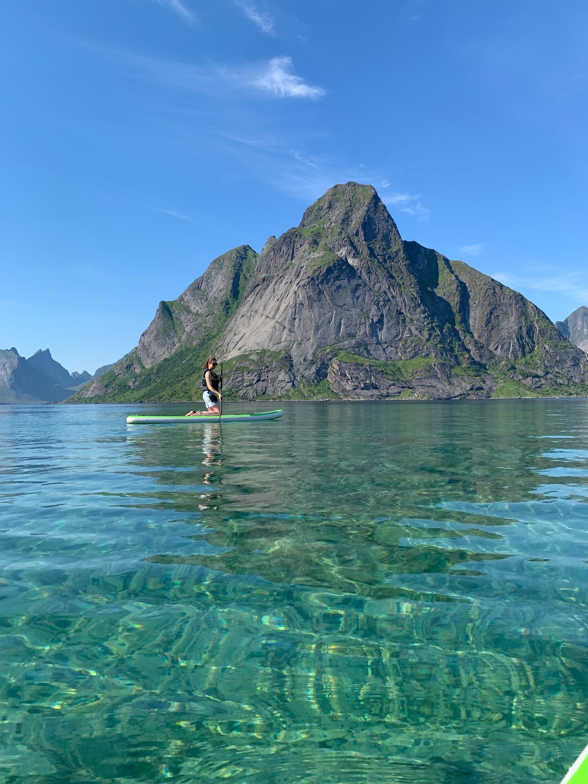 A calm body of water reflects a prominent mountain peak in the background. A stand-up paddleboarder is seen gliding across the clear water, surrounded by stunning natural scenery under a bright blue sky.