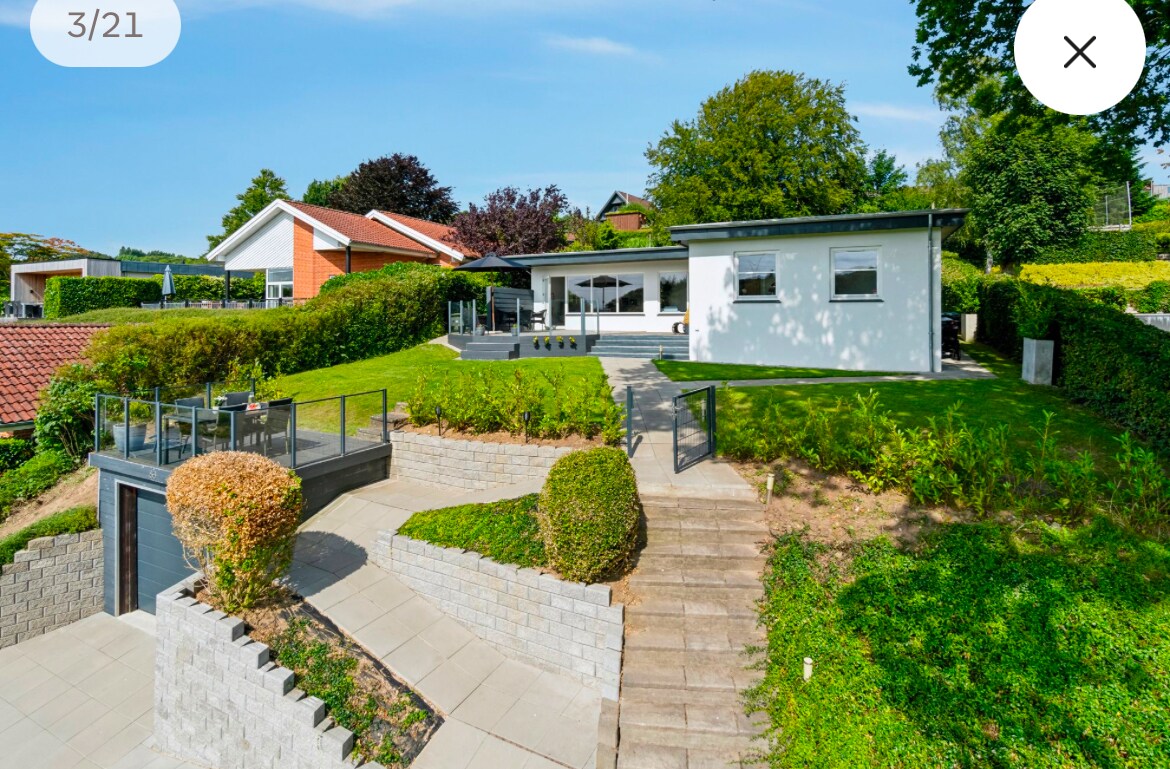 The exterior of the villa is showcased, featuring a well-maintained garden with greenery and stone pathways. The modern structure has large windows that invite natural light. A deck area with seating is visible, surrounded by lush landscaping. The serene atmosphere complements the surrounding nature.