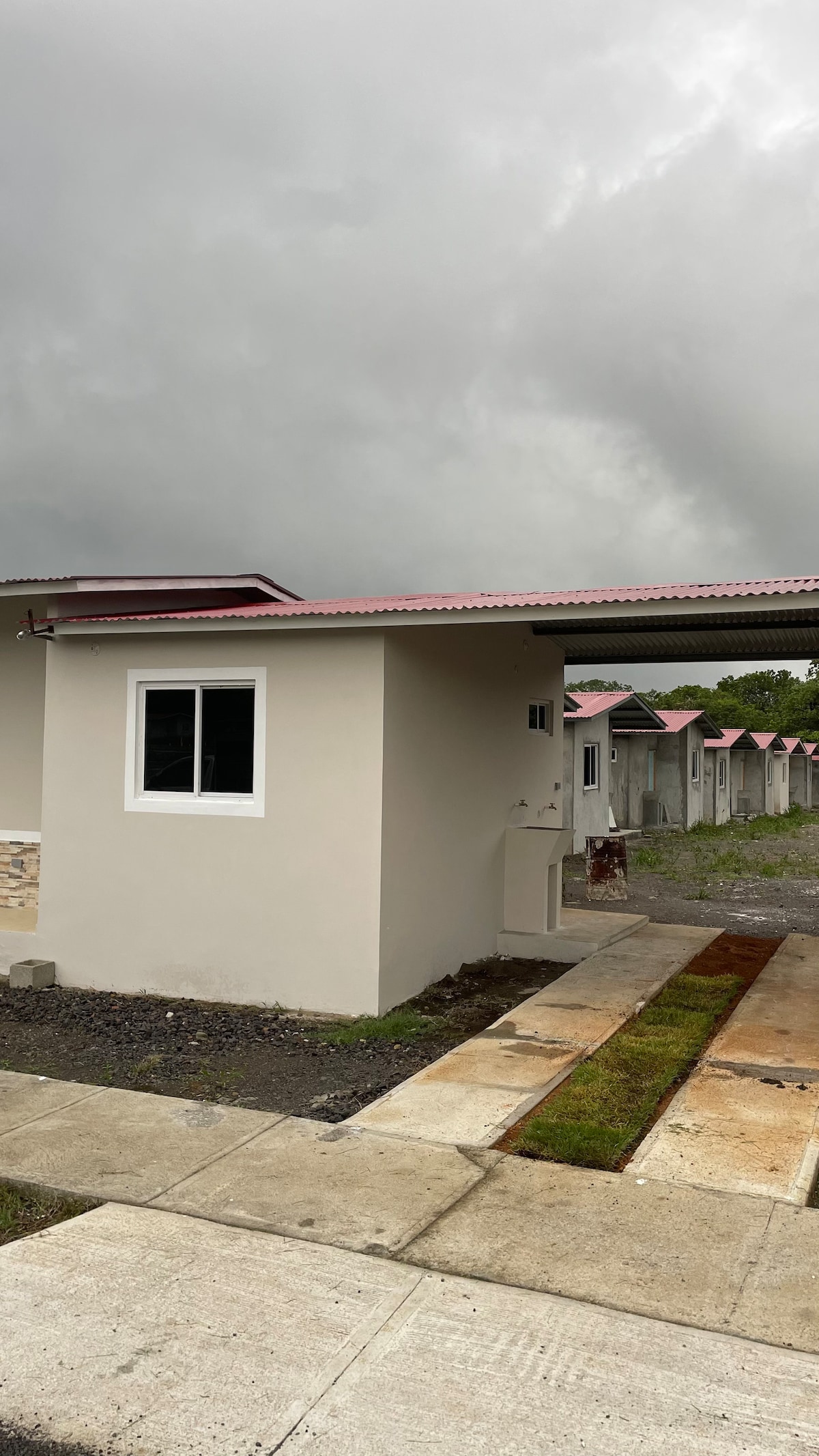 A single-story building is shown with a light-colored exterior and a red roof. A small window allows natural light inside. The surrounding pathway features stone pavers and patches of grass, leading to additional structures in the background beneath an overcast sky.